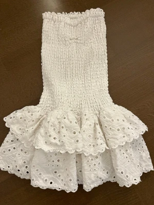 Vit kort klänning från Design by Amor - Supersöt vit kort klänning från Design by Amor med smockad överdel och volangig kjol i broderad bomull. Klänningen har fina spetsdetaljer och en romantisk känsla, perfekt för sommarens alla tillfällen.