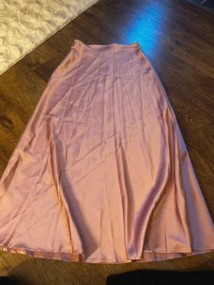 Ljusrosa satinlångkjol - Säljer en elegant ljusrosa långkjol i satin med mjuk glansig yta och enkel, stilren design. Kjolen har en lätt utställd form och faller snyggt längs benen. Perfekt för dig som gillar en feminin och trendig look.