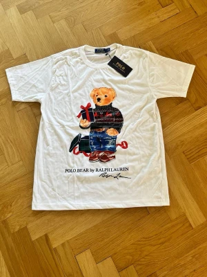 Vit Polo Bear t-shirt från Ralph Lauren - Vit t-shirt från Ralph Lauren med ikoniskt Polo Bear-motiv på bröstet. Björnen har en julig tröja och håller ett paket, vilket ger en festlig vibe. Klassisk passform och rund hals. Perfekt för dig som vill sticka ut med en stilren och lekfull look. Denna är i måtten= Axel till axel 43cm och Axel ner till botten 63cm jag försöker skicka samma dag och skriv till mig om ni vill göra bundle med den andra t shirten som ligger uppe fast i svart och storlek L👍😊
