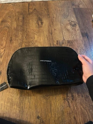 Svart necessär i krokopräglat material - Snygg svart necessär från Lyko Lovables i glansigt krokopräglat material. Perfekt för att förvara smink, hudvård eller andra småprylar. Stängs med dragkedja och har en stilren look med diskret logga på framsidan.