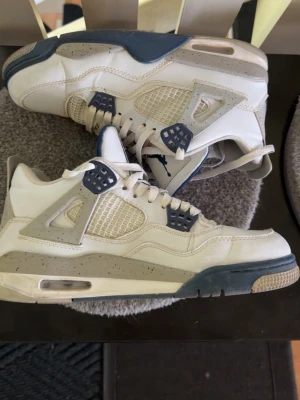 Jordan 4 - Säljer ett par vita sneakers med blå och grå detaljer. Skorna har meshpaneler på sidorna, synlig luftdämpning i sulan och specklade detaljer på mellansulan. Klassisk siluett med lågt skaft och snörning framtill. Perfekta för dig som gillar streetwear.