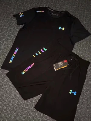 Svart träningsset från Under Armour - Säljer ett par svart träningsset från Under Armour med färgglada loggor och detaljer. Shortsen har snörning i midjan och är tillverkade i ett lätt och sportigt material som passar perfekt för träning eller chill. Stilen är modern och loggorna ger en extra färgklick. Storlek S, helt ny