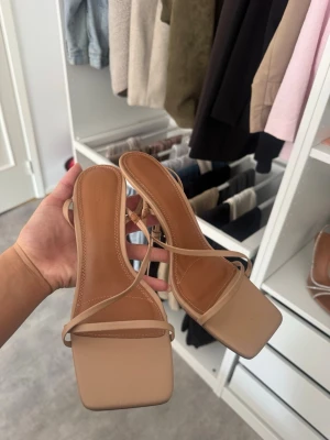 Beige sandaler med smala remmar NA-KD - Säljer ett par stilrena beige sandaler från NA-KD med smala remmar och fyrkantig tå. Sandalerna har en låg klack och är tillverkade i konstläder med snygga sömdetaljer. Perfekta för en trendig och minimalistisk look.