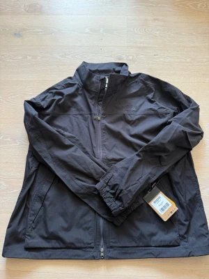 Svart vindjacka från The North Face - Snygg svart vindjacka från The North Face med dragkedja framtill, hög krage och två sidofickor. Jackan har diskret rutmönster i tyget och elastiska muddar vid ärmsluten. Perfekt för blåsiga dagar och enkel att matcha med olika outfits. Helt ny!