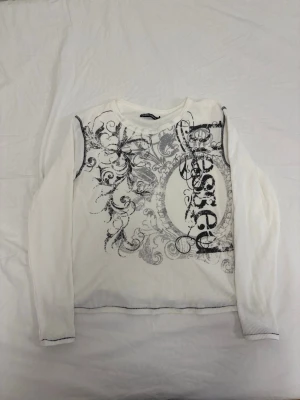 Vit långärmad topp från Insomnia Visuals - Säljer en vit långärmad topp från Insomnia Visuals med svart och grått grafiskt tryck och swirl-mönster framtill. Toppen har rund halsringning och är gjord i ett ribbat, stretchigt material som känns bekvämt och trendigt. Storlek M men passar även S