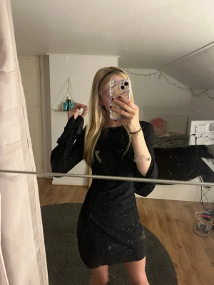 Svart glittrig kortklänning - Snygg svart kortklänning med glittriga detaljer och öppen rygg. Klänningen har lång ärm och en figurnära passform som framhäver siluetten. Perfekt för dig som vill sticka ut med lite extra glitter.