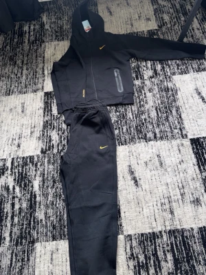 Svart Nike NOCTA loungewear set - Snyggt svart loungewear set från Nike NOCTA med gul logga på både hoodie och byxor. Hoodien har dragkedja, huva och fickor med dragkedja. Byxorna har resår i midjan och praktiska fickor. Perfekt för chill dagar hemma eller när du vill vara bekväm.