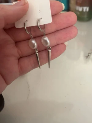 Silvriga örhängen med pärlor och spikes - Säljer ett par trendiga örhängen i silverfärg med vita pärlor och spetsiga spikes. Örhängena har en cool kombination av klassiska pärlor och edgy metallspetsar som ger en unik look. Perfekta för dig som vill sticka ut lite extra.