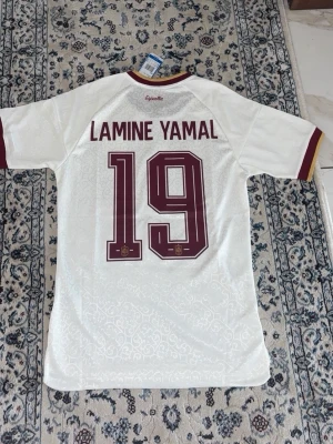 Vit Spanien Lamine Yamal matchtröja - Säljer en vit Spanien matchtröja från Adidas med Lamine Yamal och nummer 19 på ryggen. Tröjan har vinröda detaljer, korta ärmar och officiella märken på bröstet. Tillverkad i ett lätt och ventilerande material, perfekt för fotboll eller träning.