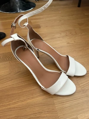 Vita klackskor med ankelrem - Stilrena vita klackskor med öppen tå och bred rem över foten. Skorna har en låg klack och justerbar ankelrem med spänne. Tillverkade i ett slätt material med beige innersula för extra komfort. Perfekta för att lyfta din outfit.