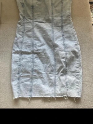 Ljusblå axelbandslös jeansklänning - Säljer en ljusblå, axelbandslös jeansklänning med markerade sömmar och rå kant nertill. Klänningen har en tight passform och dragkedja bak. Perfekt för dig som gillar en edgy och trendig look.