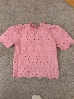 Rosa broderad blus med puffärm - Söt rosa blus med broderade hål och puffiga korta ärmar. Blusen har volangkant vid halsen och nederkanten samt en liten knapp i nacken. Materialet är skönt mot huden