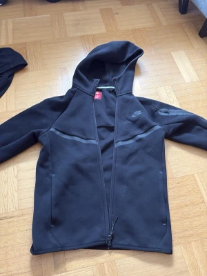 Svart Nike hoodie med dragkedja - Säljer en svart hoodie från Nike med hel dragkedja framtill och huva. Hoodien har två sidofickor och en liten ficka på ärmen. Tillverkad i ett mjukt och stretchigt material, perfekt för en sportig och avslappnad stil. Diskret Nike-logga på bröstet.