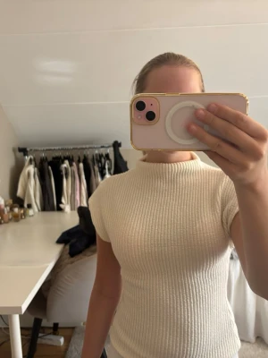 Ribbad vit topp med kort ärm - Säljer en ribbad vit topp med korta ärmar och hög hals. Toppen har en slim passform och är tillverkad i ett mjukt, stretchigt material som sitter snyggt på kroppen. Perfekt att styla med jeans eller kjol för en clean look.
