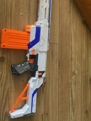 Nerf gevär med orange magasin - Säljer ett coolt Nerf gevär i vitt, blått och orange med svart handtag. Geväret har ett avtagbart orange magasin och detaljer i plast. Perfekt för lek och actionfyllda Nerf-strider med kompisarna!