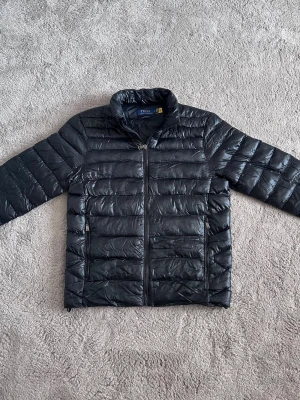 Svart dunjacka från Polo Ralph Lauren - Snygg svart dunjacka från Polo Ralph Lauren med diskret logga på bröstet. Jackan har hög krage, två sidofickor med dragkedja och quiltad design. Perfekt för kalla dagar och tillverkad i ett glansigt material som ger en modern look.