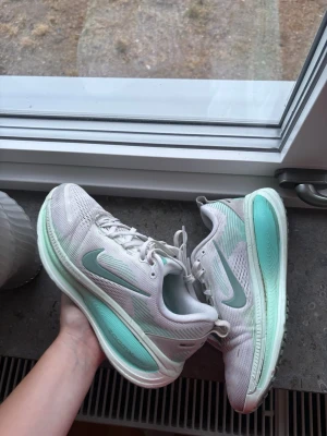 Nike vita och mintgröna löparskor - Säljer ett par snygga Nike Vomero 18 löparskor i vitt med mintgröna detaljer och swoosh. Skorna har en tjock sula med ZoomX-teknologi för extra komfort och respons. Ovandelen är i meshmaterial som andas och har klassisk snörning. Perfekta för träning eller promenader.