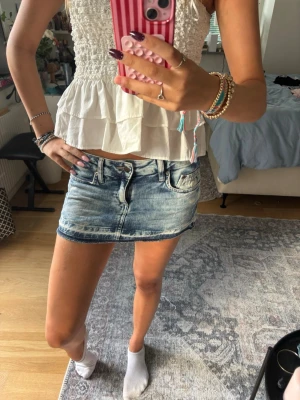 Blå jeansminikjol med slitningar - Trendig blå jeansminikjol med slitna detaljer och klassiska fickor fram och bak. Kjolen har en låg midja och rå kant nedtill för en avslappnad look. Perfekt att matcha med crop tops eller hoodies för en cool vardagsstil.
