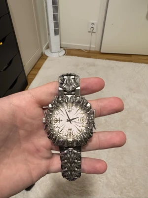 Seams klocka  - Säljer en riktigt unik klocka i silverfärg med detaljerat, nästan gotiskt mönster på både boett och armband. Urtavlan är vit med dekorativa svarta visare och ornament. Perfekt för dig som vill sticka ut med accessoarerna. Nypris 4700kr