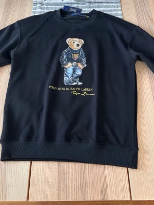 Svart Polo Bear hoodie Ralph Lauren - Svart hoodie från Ralph Lauren med ikoniskt Polo Bear-motiv på bröstet. Trycket visar en björn i jeans och skinnjacka. Klassisk rund halsringning och ribbade muddar. Perfekt för en avslappnad och trendig stil.