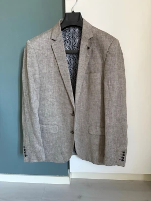 Ljusgrå linnékavaj slim fit - Stilren ljusgrå kavaj i linnemix från Dressmann med slim fit-passform. Kavajen har två knappar framtill, fickor med lock och dekorativa knappar vid ärmslut. Insidan har ett paisleymönstrat foder och randiga detaljer för en extra snygg touch.
