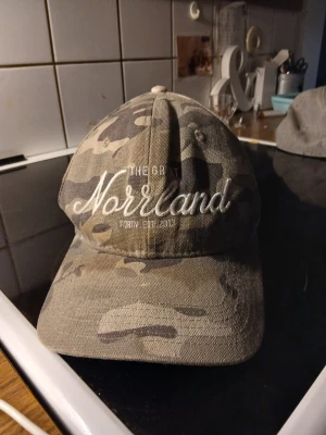 Camo keps med Norrland-brodyr - Snygg keps i camomönster med broderad text 'Norrland' framtill. Klassisk böjd skärm och justerbar baksida. Perfekt för dig som gillar outdoor-stil och vill visa kärlek till Norrland.