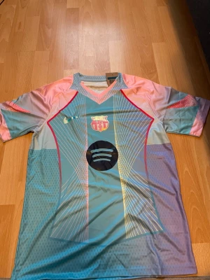 FC Barcelona matchtröja i pastell - Säljer en FC Barcelona matchtröja i pastellfärger med Nike-logga och Spotify-logga på bröstet. Tröjan har korta ärmar, V-ringning och ett unikt mönster i blått, rosa och lila. Perfekt för fotbollsträning eller att bära som supporter.
