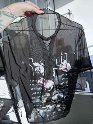 Svart mesh topp med snörning MISSGUIDED - Säljer en svart mesh topp med snörning framtill och tryck med lila rosor och text. Toppen är kortärmad och har en edgy vibe med transparent material och coola detaljer. Perfekt för dig som vill sticka ut och gillar streetstyle. Aldrig använd, endast testad 