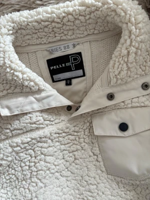 Vit teddytröja från Pelle P - Mysig vit teddytröja från Pelle P med hög krage och knappdetaljer. Tröjan har en bröstficka med lock och svarta knappar samt ribbade muddar. Perfekt för kalla dagar och riktigt skön att bära. Materialet är fluffigt och värmande.