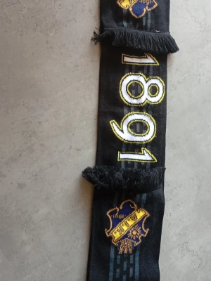 Svart AIK halsduk med brodyr - Svart halsduk med broderad AIK-logga och årtalet 1891 i gult och vitt. Halsduken har fransar i ändarna och detaljer i blått och gult. Perfekt för dig som vill visa ditt AIK-stöd.