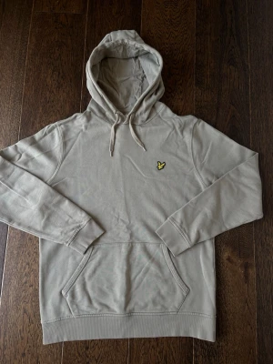 Grön Lyle & Scott hoodie - Snygg grön hoodie med huva och dragsko. Har en gul broderad logga på bröstet och en klassisk känguruficka framtill. Perfekt för en avslappnad och trendig stil. Mjukt material och ribbade muddar vid ärmslut och nederkant.