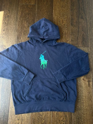 Ralph Lauren hoodie med grön logga - Snygg mörkblå hoodie från Polo Ralph Lauren med stor grön logga på bröstet. Klassisk känguruficka framtill och huva. Perfekt för en avslappnad och trendig look. Mjuk bomullskvalitet och ribbade muddar vid ärmar och nederkant.