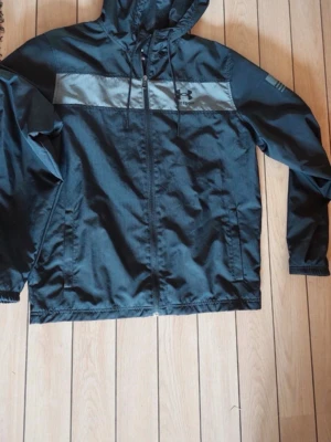 Svart vindjacka från Under Armour - Snygg svart vindjacka från Under Armour med grått bröstparti och logga framtill. Jackan har huva med dragsko, hel dragkedja och två sidofickor. Perfekt för blåsiga dagar och sportiga outfits.