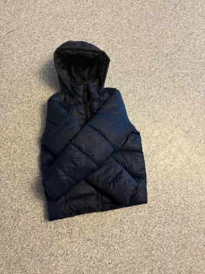 Mörkblå dunväst Wayfarer Young - Snygg mörkblå dunväst från Wayfarer Young med huva och breda, quiltade paneler. Västen har dragkedja framtill och är perfekt för lager på lager under kyliga dagar. Materialet är glansigt och ger en modern look.