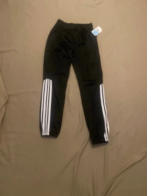 Svarta Adidas träningsbyxor - Svarta träningsbyxor från Adidas med klassiska vita ränder längs benen. Byxorna har resår i midjan och vid bensluten, samt en sportig passform som passar perfekt till träning eller chill. Materialet är lätt och andas bra.