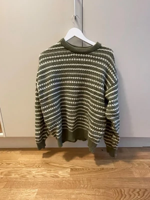 Olivgrön stickad tröja med ränder - Snygg olivgrön stickad tröja med vita och mörkgröna horisontella ränder. Tröjan har rund halsringning, ribbade muddar och avslut. Perfekt för lager-på-lager och chill stil. Materialet känns mjukt och mysigt.
