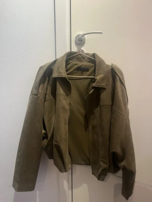 Olivgrön mockajacka med krage - Snygg olivgrön mockajacka med klassisk krage och raka ärmar. Jackan har en enkel design utan synliga detaljer på framsidan och är tillverkad i mjukt skinnmaterial. Perfekt för dig som vill ha en stilren jacka med avslappnad passform.