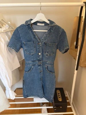 Jeansklänning med kort ärm och knappar - Snygg jeansklänning i blå denim med kort ärm, v-ringning och knappar framtill. Klänningen har två bröstfickor och två fickor vid höfterna, samt markerad midja för en smickrande passform. Perfekt för en trendig och avslappnad look.