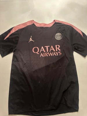 Svart PSG x Jordan matchtröja - Säljer en svart PSG x Jordan matchtröja med rosa detaljer och logga på bröstet. Tröjan har korta ärmar, rund hals och är tillverkad i ett lätt och ventilerande material. Qatar Airways tryck på framsidan. Perfekt för fotboll eller träning.
