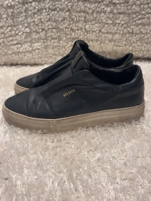 Svarta sneakers från Axel Arigato - Stilrena svarta sneakers från Axel Arigato i slätt läder med diskret guldfärgad logga på sidan. Skorna har en chunky vit sula och minimalistisk design utan synliga snören, vilket ger en clean look. Perfekta för dig som gillar enkel och modern stil. Skriv för mer info