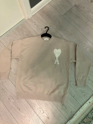 Beige stickad tröja från Ami - Snygg beige stickad tröja från Ami med rund halsringning och ribbade muddar. På bröstet finns ett vitt hjärta med bokstaven A, vilket ger en stilren och trendig look. Perfekt för dig som gillar minimalistisk design och mjuka material. Helt ny, inte använd.