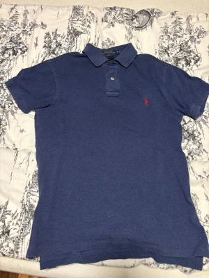 Blå pikétröja från Ralph Lauren - Klassisk marinblå pikétröja från Ralph Lauren med korta ärmar och krage. Tröjan har en liten röd broderad logga på bröstet och knäppning vid halsen. Perfekt för en stilren och avslappnad look.