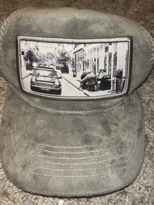 Grå meshkeps med fototryck - Unik grå keps med mjuk mockaliknande framsida och mesh på sidorna. På fronten sitter ett svartvitt fototryck av en stadsscen med bil och uteservering. Justerbar baktill för perfekt passform. Perfekt för dig som gillar streetstyle och detaljer.