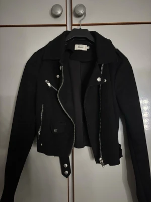 Svart biker skinnjacka från ONLY - Snygg svart skinnjacka från ONLY med klassisk bikerstil, silvriga dragkedjor, fickor och bälte i midjan. Jackan har bred krage och coola detaljer som ger en edgy look. Perfekt för dig som vill ha en stilren och trendig jacka.