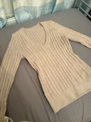 Beige kabelstickad tröja från SHEIN - Säljer en beige kabelstickad tröja från SHEIN med djup V-ringning och långa ärmar. Tröjan är tillverkad i en mjuk mix av akryl och polyester och har ribbade muddar vid ärmslut och nederkant. Perfekt för lager på lager och en clean look.