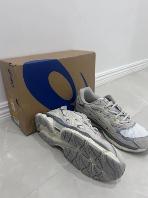 Asics grå och beige sneakers - Snygga sneakers från Asics i grått och beige med meshpaneler och detaljer i mocka och syntet. Skorna har en chunky sula och klassisk Asics-logga på sidan. Perfekta för dig som gillar sportig och trendig stil.