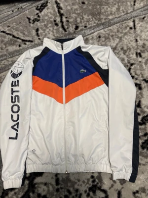 Lacoste vit vindjacka med färgblock - Snygg vindjacka från Lacoste i vitt med blå och orange färgblock på bröstet samt svarta detaljer. Dragkedja framtill, resår i ärmslut och nederkant. Stor Lacoste-logga och grafiskt tryck på ena ärmen. Klassisk krokodil-logga på bröstet. Perfekt för en sportig look.