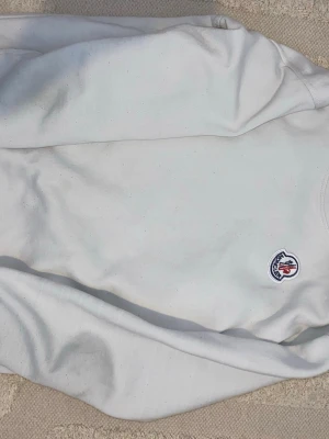 Vit sweatshirt från Moncler - Säljer en stilren vit sweatshirt från Moncler med det klassiska broderade logotypmärket på bröstet. Tröjan har rund halsringning och långa ärmar. Perfekt för dig som gillar exklusiva och enkla plagg.