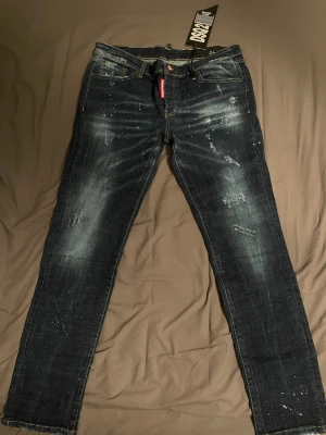 Mörkblå Dsquared2 jeans med slitningar - Säljer ett par mörkblå jeans från Dsquared2 med coola slitningar och färgstänk. Jeansen har raka ben, klassisk femficksmodell och en röd logotagg vid gylfen. Materialet är denim med snygg tvätt och detaljer som ger en trendig look.