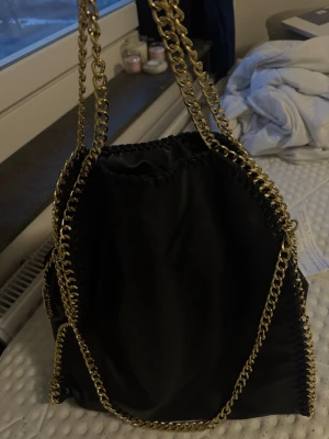 Svart axelväska med guldkedja - Säljer en svart axelväska i fejkläder med snygga guldfärgade kedjor som axelremmar och dekor längs kanterna. Väskan har en mjuk, slouchy form och cool detaljsöm runt hela väskan. Perfekt för dig som gillar statement-accessoarer.